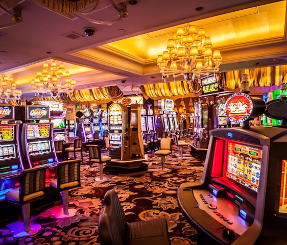 A vibrant casino interior filled with rows of colorful slot machines, each illuminated with bright lights and displays. The ceiling is adorned with elegant chandeliers and golden drapes, creating a luxurious atmosphere. The patterned carpet adds to the elaborate design of the space.