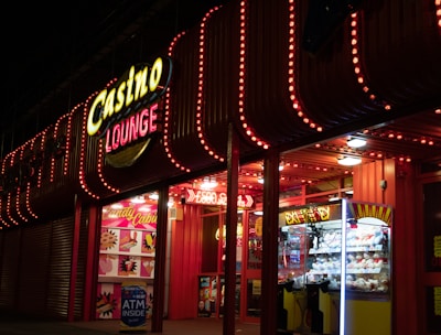 A brightly lit casino lounge entrance adorned with neon lights. The exterior features colorful signage including the words 'Casino' and 'Lounge' in vivid colors, with additional lights framing the building. Inside, a glimpse of a claw machine filled with plush toys and arcade games is visible. The area exudes an inviting yet bustling atmosphere typical of entertainment venues.